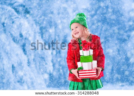 Little girl in red and green knitted hat holding Christmas present boxes in winter park on Xmas eve. Kids play outdoor in snowy winter forest. Children opening presents. Toddler kid playing with gifts