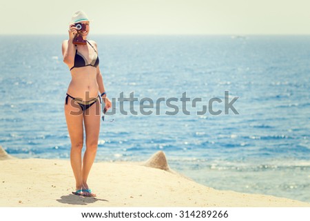 Young slim woman with old camera taking photo on the beach near the sea or ocean. Smiling girl in bikini having fun on summer vacation. Warm color toned image
