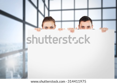 Business partners hiding behind a sign against room with large windows showing city