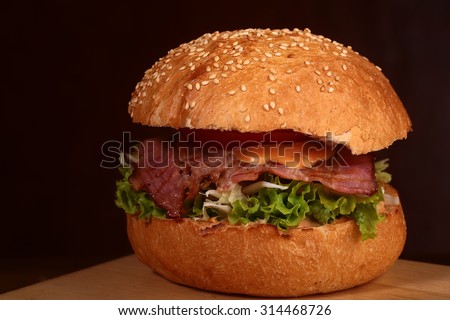 One big delicious fresh burger with green lettuce red tomato cheese cabbage bacon slice meat cutlet and white bread bun with sesame seeds on black background closeup, horizontal picture