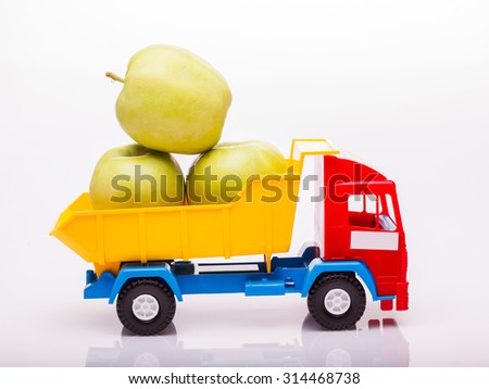 Small colorful toy truck yellow blue and red colors with three fresh green apple fruits in basket on white studio background closeup, horizontal picture