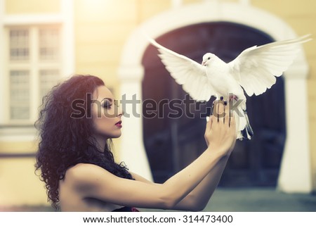 Charming beautiful delicate young woman with bright make-up with long curly brown hair in profile holding a white pigeon in her hands on blur building background outdoor, horizontal picture 