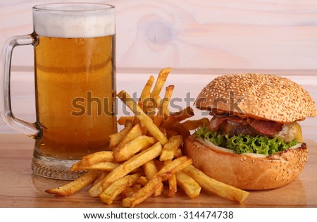 Big fresh tasty burger of green lettuce meat cutlet cheese tomato and white bread bun with sesame seeds near chips and glass of light beer on octoberfest holiday, horizontal picture