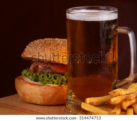 Big fresh tasty burger of green lettuce meat cutlet cheese tomato and white bread bun with sesame seeds near chips and glass of dark beer on octoberfest holiday, square picture