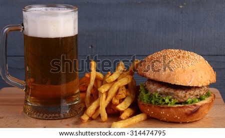 Big fresh tasty burger of green lettuce meat cutlet cheese tomato and white bread bun with sesame seeds near chips and glass of light beer on octoberfest holiday on grey background, horizontal picture