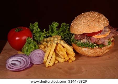 One big tasty appetizing fresh burger of green lettuce red tomato cheese bacon slice meat cutlet violet oinion white bread bun with sesame seeds and chips on wooden table closeup, horizontal picture