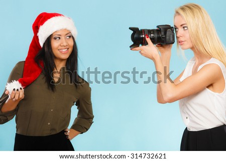 Photographer and model. Caucasian blonde girl shooting images, taking photos with camera., photographing fashion model mixed race woman in santa helper hat.