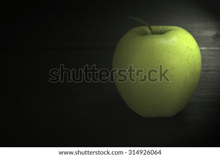 Alone green apple is standing on a wooden table. Edited as a rustic photo with dark background. Soft light illuminates the apple from the top right corner of the photo.