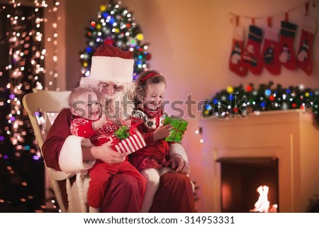 Santa Claus and children opening presents at fireplace. Kids and father in Santa costume and beard open Christmas gifts. Little girl helping with present sack. Family under Xmas tree at fire place.