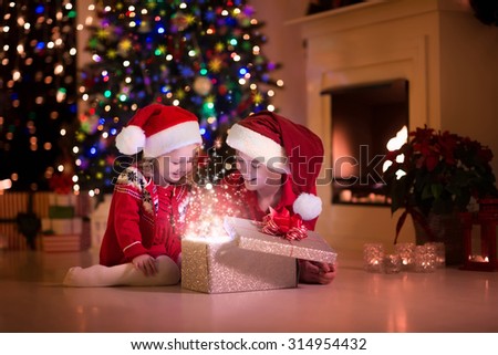 Family on Christmas eve at fireplace. Kids opening Xmas presents. Children under Christmas tree with gift boxes. Decorated living room with traditional fire place. Cozy warm winter evening at home.