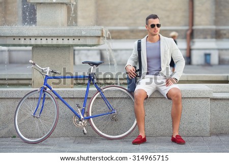 Handsome young man sitting near the fountain with his bicycle beside him
