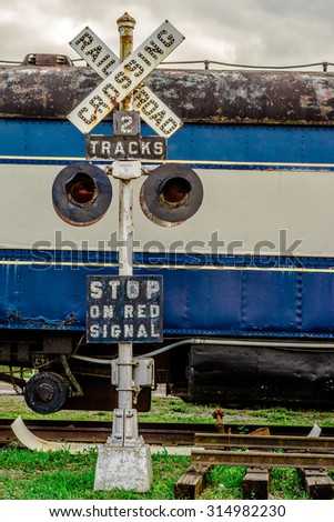 A Damaged Decaying Railroad crossing sign in front of an abandoned Locomotive on a set of broken railroad tracks.