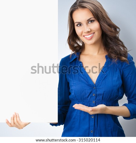 Portrait of happy smiling young woman in blue clothing, showing blank signboard with blank copyspace area for slogan or text, posing at studio