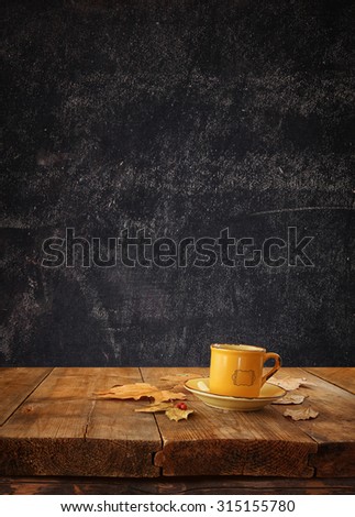 front image of coffee cup over wooden table and autumn leaves in front  and blackboard background with room for text