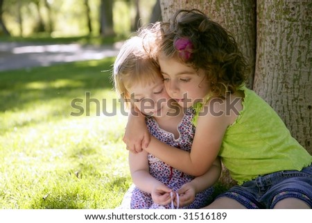 Two little sister girls hug playing under a tree trunk in the park