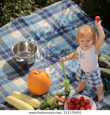 One little cook smiling boy at picnic standing with ladle pot orange pumpkin squash and cucumber holding red tomato with food sitting on checkered plaid on natural background sunny day, square picture