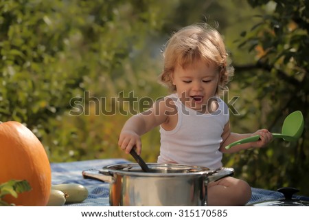 Closeup of one funny cook boy at picnic sitting with black ladle pot orange pumpkin and other vegetables playing with food sitting on blue checkered plaid on natural background, horizontal picture