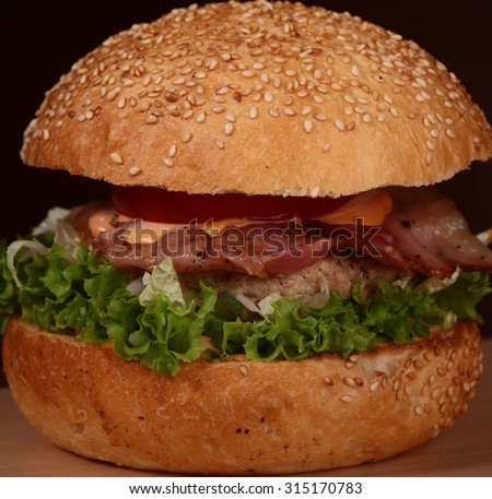 One big delicious appetizing fresh burger of green lettuce red tomato cheese and bacon slice and white bread bun with sesame seeds on bkack background closeup, square picture