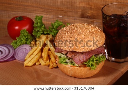 Big tasty appetizing fresh burger of green lettuce red tomato cheese bacon slice meat cutlet violet oinion and white bread bun with sesame seeds and chips with glass of cold cola, horizontal picture