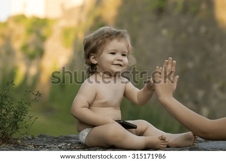 One little curious smiling baby boy with blonde curly hair and cute face holding and playing on black mobile phone touching female hand sitting outdoor on natural background, horizontal picture