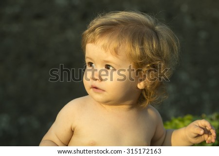 Closeup of little smiling interested cute baby boy with blonde curly hair round cheeks playful eyes standing without clothes outdoor lookling away on natural background, horizontal picture