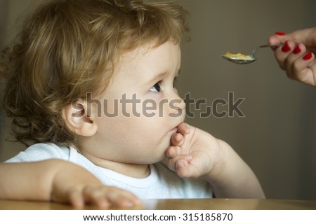 Sad frown serious cute charming little boy child with brown hair and hazel eyes looking at spoon holding by mother hand and cover mouth by his palm on grey background indoor, horizontal picture