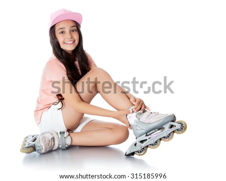 Charming dark-haired girl of school age in short white shorts and a pink t-shirt sitting on the floor and tries to foot roller skates. -Isolated on white background