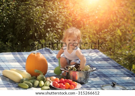 One little cook boy at picnic sitting with ladle pot orange pumpkin red tomato squash and cucumber playing with food sitting on blue checkered plaid on natural background sunny day, horizontal picture