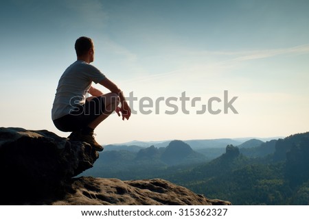 Young hiker in black pants and shirt  is sitting on cliff's edge and looking to misty hilly valley bellow. Enjoy life.Hiker sitting. Outdoor man enjoy.Cliff edge. Looking over horizon edge. Hike man. Royalty-Free Stock Photo #315362327