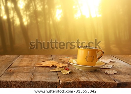 front image of coffee cup over wooden table and autumn leaves in front of autumnal sunset background
