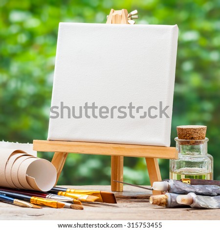A blank canvas on easel, artistic paintbrushes, tubes of oil paint and bottle of flax seed oil.