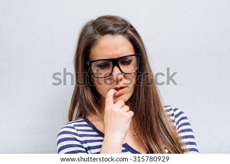 Young woman in glasses thinking, finger to her mouth. On a gray background.