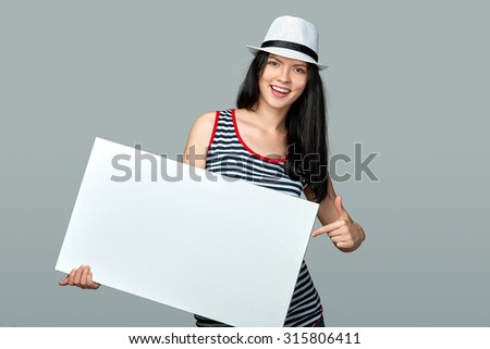 Smiling woman pointing on white empty billboard over grey background in studio.