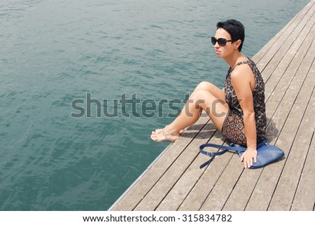 Portrait of beautiful stylish woman relaxing after active walking while sitting on a wooden pier with copy space in a summer day, young female tourist enjoying rest and good weather during her weekend
