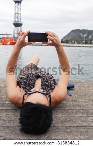 Brunette woman holding cell telephone with copy space area for your text message or advertising content, female making self portrait with her smart phone digital camera while lying on a pier