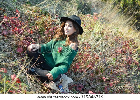 Picture of young girl in black hat and sweater sitting on dried grass with read leaves. Pretty woman smiling on sunny autumn outdoor background.
