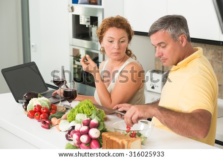 Serious woman looking at her husband preparing vegetables salad. Mature man cutting cucumbers and tomatoes in the kitchen.