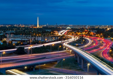 Washington D.C. cityscape at dusk with rush hour traffic trails on I-395 highway. Washington Monument, illuminated, dominates the skyline.