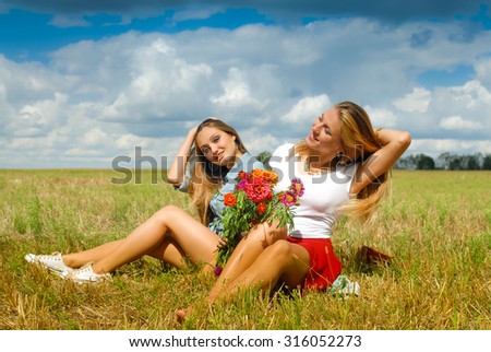 Picture of two pretty girls sitting on grass field with red flowers. Young women smiling and enjoying summer on blue sky outdoor blackground.