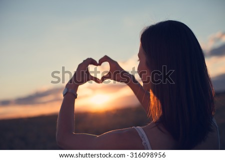 A young girl making heart symbol with her hands at sunset (lens focus on face) Royalty-Free Stock Photo #316098956