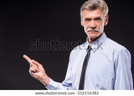 Senior man in shirt is pointing with finger away, dark background.