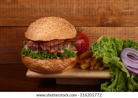 One big tasty appetizing fresh burger of green lettuce red tomato cheese bacon slice meat cutlet violet oinion and white bread bun with sesame seeds on wooden background closeup, horizontal picture