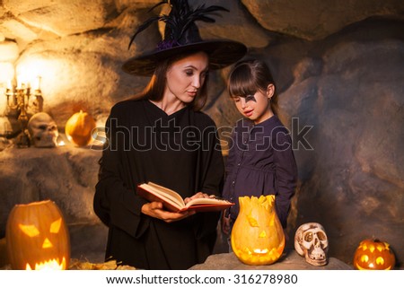 Attractive young witch is holding a book of spells. She is showing it to a little girl and smiling slyly. The kid is looking at it with interest. They are standing in cave and celebrating Halloween