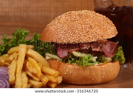 Big tasty appetizing fresh burger of green lettuce red tomato cheese bacon slice meat cutlet violet onion and white bread bun with sesame seeds and chips with glass of cold cola, horizontal picture