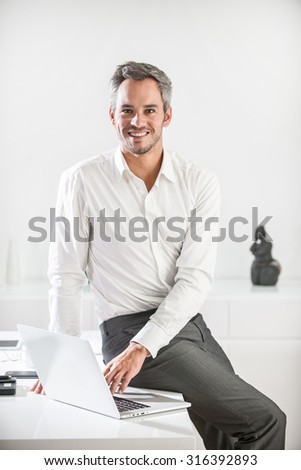 Portrait of a grey hair businessman with beard sitting on his desk in a luminous office. He is wearing a grey suit pant and a white shirt, he is looking at camera, his laptop in front of him
