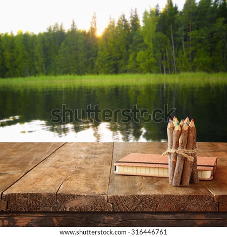 vintage notebook and stack of wooden colorful pencils on wooden texture table in front of countryside lake and forest view 