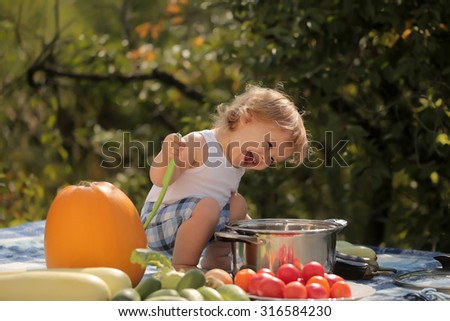 One playful boy at picnic sitting with ladle pot orange pumpkin red tomato squash and cucumber playing with food sitting on blue checkered plaid on natural background sunny day, horizontal picture