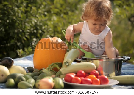 Cute small child at picnic holding ladle sitting near pot among orange pumpkin red tomato squash and cucumber on blue checkered plaid on natural background sunny day, horizontal picture