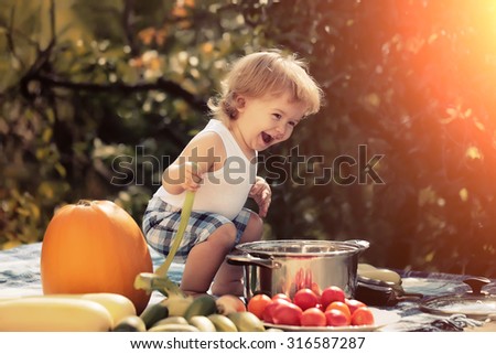 One playing boy at picnic sitting with ladle pot orange pumpkin red tomato squash and cucumber playing with food sitting on blue checkered plaid on natural background sunny day, horizontal picture