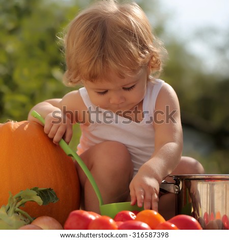 Cute little boy at picnic holding ladle sitting near pot among orange pumpkin red tomato squash and cucumber on blue checkered plaid on natural background sunny day, square picture